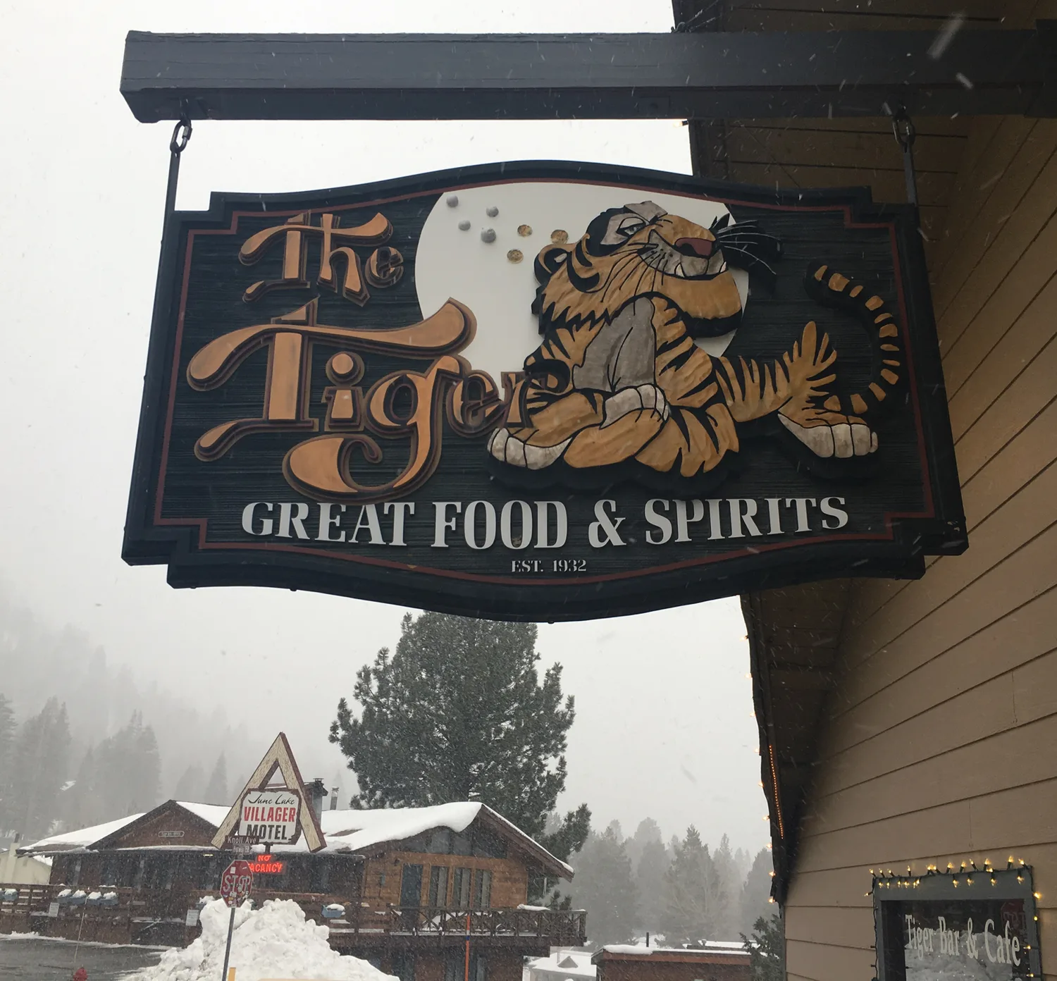 The carved Tiger Bar sign hanging off the building in falling snow: a painted tiger on a black board, THE TIGER in amber letters, GREAT FOOD & SPIRITS and EST. 1932 beneath it