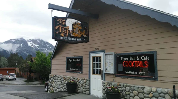 The Tiger Bar & Cafe building on Highway 158: tan clapboard, the hanging tiger sign, COCKTAILS and Established 1932 lettered on the window, a snowy Sierra ridge at the end of the street